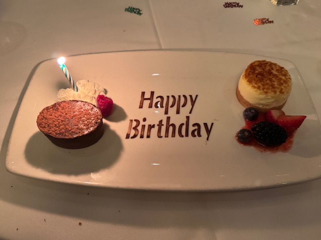 Assiette de dessert avec "Happy Birthday" dessiné en poudre de cacao au centre, un petit cheesecake, et un petit gâteau au chocolat sans farine.