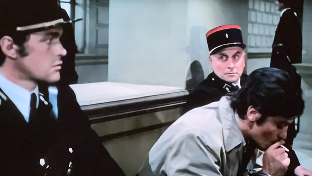 Delon fume sur un banc, entre deux gendarmes