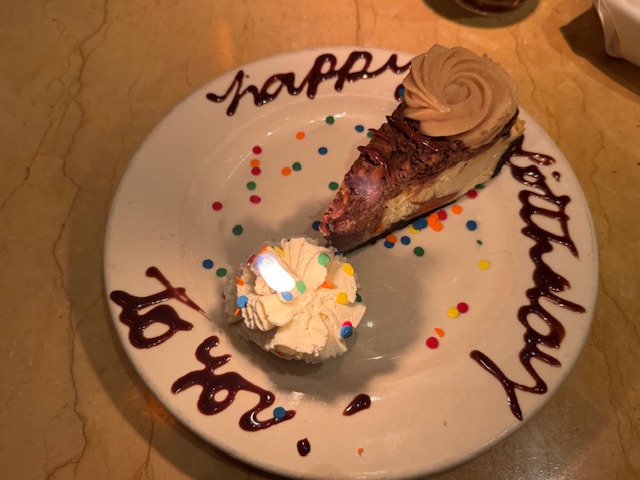 Assiette avec un bout de cheesecake, une bougie allumée et les mots "Happy Birthday" écrits en chocolat