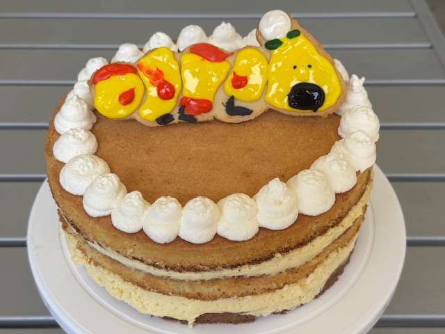 Gâteau Wiggler : un fond de biscuit chocolat-amande, avec deux couches de mousse au chocolat blanc et deux couches de génoise en haut. Il y a des sphères de crème Chantilly autour des bords en haut, et au centre, un biscuit en pâte sucrée est coupé en forme de chenille Wiggler, glacé en jaune, avec des pois oranges, un nez noir, une fleur blanche et verte sur la tête, et des chaussures violettes.