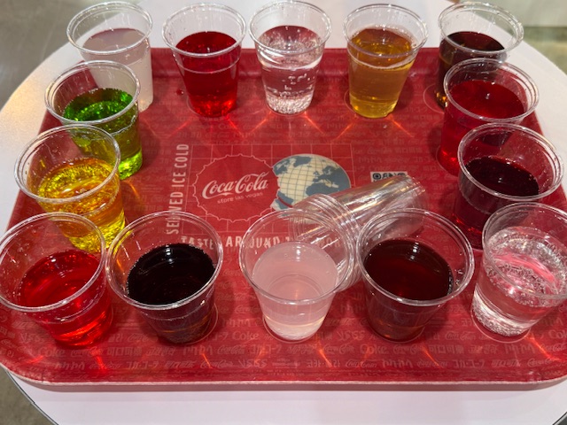 14 petits verres en plastique avec des sodas ; les couleurs sont toutes très artificielles