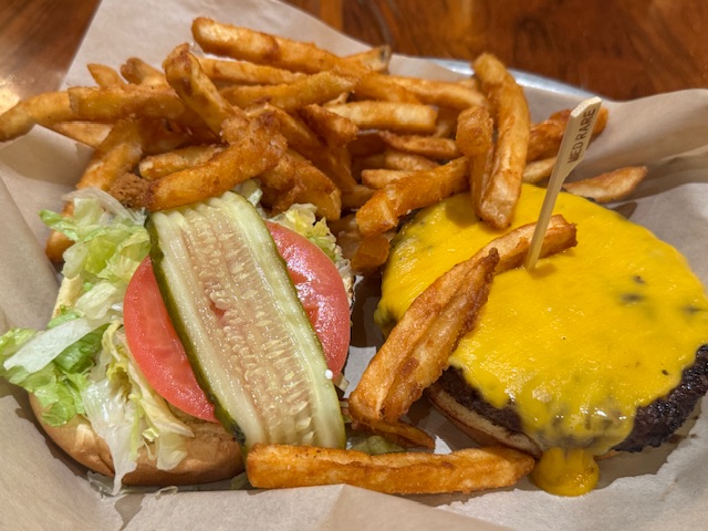 Photo de hamburger : à gauche, la moitié du pain est recouvert de laitue, d'une tranche de tomate, et d'un cornichon. À droite, l'autre moitié du pain contient la galette de viande ainsi que du fromage cheddar bien jaune. Il y des frites en haut.
