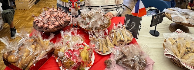 Photo beaucoup plus large que haute avec, de gauche à droite : des financiers, des macarons, des madeleines aux amandes, des nougatines, des biscuits en forme de Tour Eiffel, et des chocolats. Un panneau derrière les biscuits de Péla indique que toute la table est dispo pour 2 $ la pièce.
