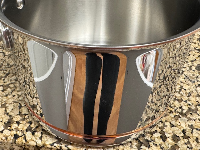 Casserole en inox sur un comptoir en faux marbre. La surface est très polie, alors on peut voir un reflet déformé de mon pantalon, ainsi que les portes des placards derrière moi. Il y a une bande de cuivre juste au-dessus du fond.