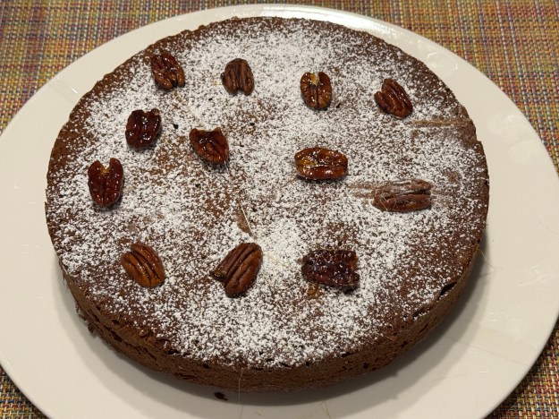 Gateau entier sur une assiette -- le gâteau a une couleur marron foncé, c'est parfaitement rond et fait 23 cm de large, il y a du sucre glace tamisé partout, et une douzaine de noix de pécan caramelisées parsemées au-dessus