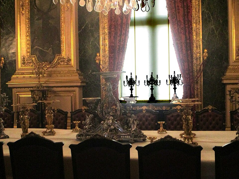C'est une salle à manger avec un surtout de table en métal argenté de Christofle. Les murs sont dorés comme on trouve souvent dans les palais, Les sièges sont en cuir et en bois, et il y a trois candélabre à 10 branches devant la fenêtre, elle même recouverte d'un rideau blanc. 