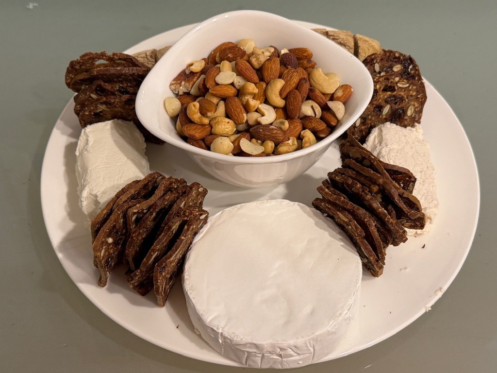 Plateau d'apéro : baguette largement cachée derrière un bol de fruits secs, deux fromages de chèvre, un rond de brie, et 4 tas des biscuits "Raincoast Crisps" qui séparent les fromages.