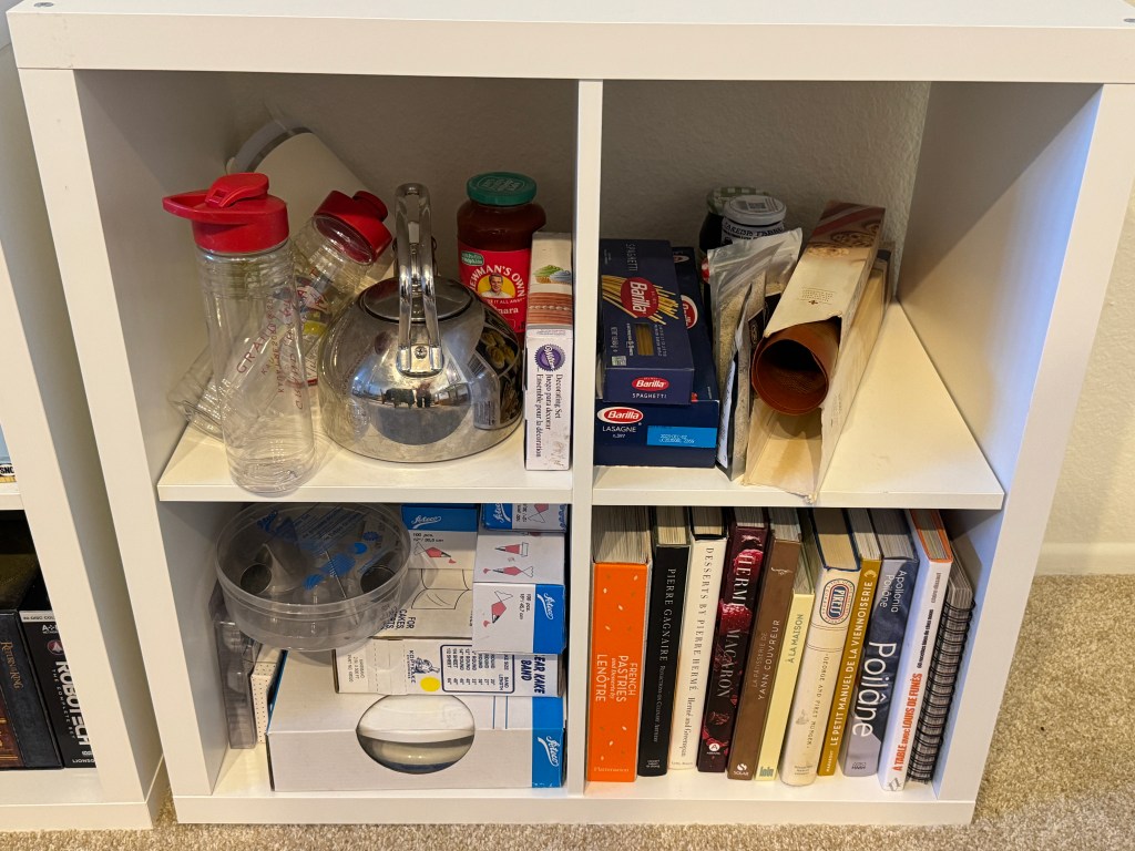 Bibliothèque à 4 étagères en forme de cube. En haut à gauche, une théière et des bouteilles d'eau ; à droite, des boites de pâtes, mes tapis en silicone et de gros sacs de sucre vanillé. En bas à gauche, des poches à douille ; en bas à droite, ma collection de livres de cuisine en français.