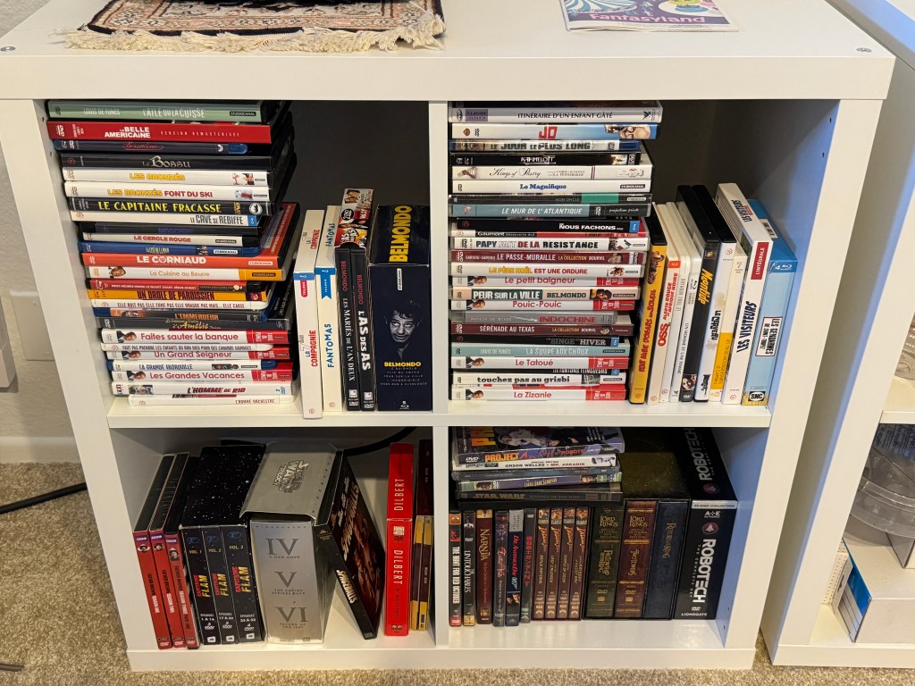 Bibliothèque en forme de cube, avec 4 étagères au total. Sur les 2 en haut, il n'y a que des DVDs et des Blu-Rays français, dont une vingtaine de films de Louis de Funès ainsi que des coffrets de Fantômas, de la 7e Compagnie, des Gendarmes de Saint-Tropez et de Belmondo. En bas, il y a des disques anglophones -- Father Brown de la BBC, Star Wars, le Seigneur des Anneaux, Indiana Jones et Matrix.