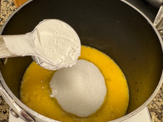 Casserole avec des œufs battus là-dedans et un tas de sucre au centre. Une cuillère pleine de maïzena se tient au-dessus de la casserole