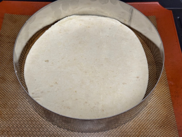 Disque de pâte visiblement sur un tapis en silicone, avec un cercle de pâtisserie autour des bords, pour voir que c'est maintentant un peu en forme d'ovale