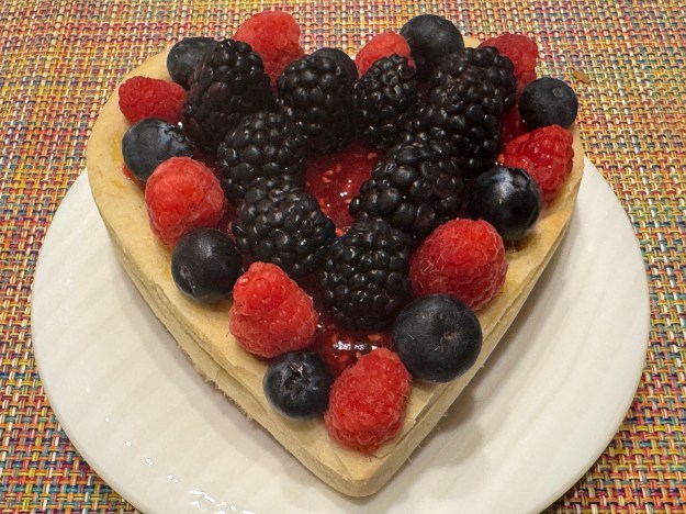 C'est une tartelette en forme de cœur sur une assiette juste un peu plus large que la tartelette (qui fait environ 13 cm de large). Il y a une bande de framboises et de myrtilles aux bords, puis des mûres à l'intérieur, et juste un petit cercle de confiture de framboises visible au centre