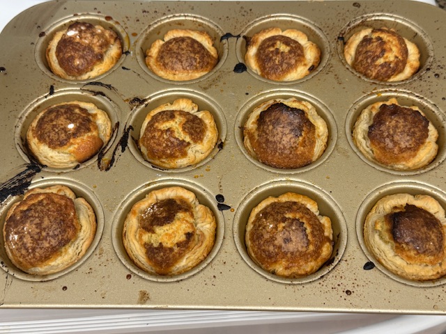 Mirlitons sortis du four, toujours dans le moule. Il y a des taches noirs où le sucre glace a brûlé sur le mêtal. Les centres sont bien gonflés et dorés.