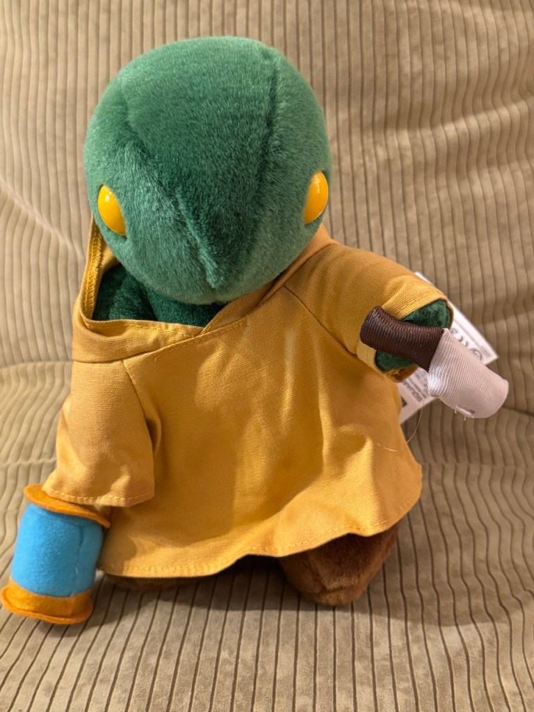 Tomberry vu d'en face : il est vert et ressemble à un lézard, s'il marchait sur deux pieds, avec deux yeux jaunes aux côtés de la tête. Il porte une soutane marron comme un moine, où la capote est derrière sa tête. Dans la main droite, il porte une lanterne, et dans la main gauche, un gros couteau (tout en peluche).