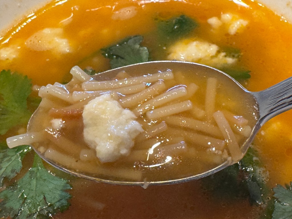Cuillère de soupe de fideo en gros-plan, avec du fromage et des nouilles clairement visibles. Le bol de soupe, avec des feuilles de coriandre, est en arrière-plan