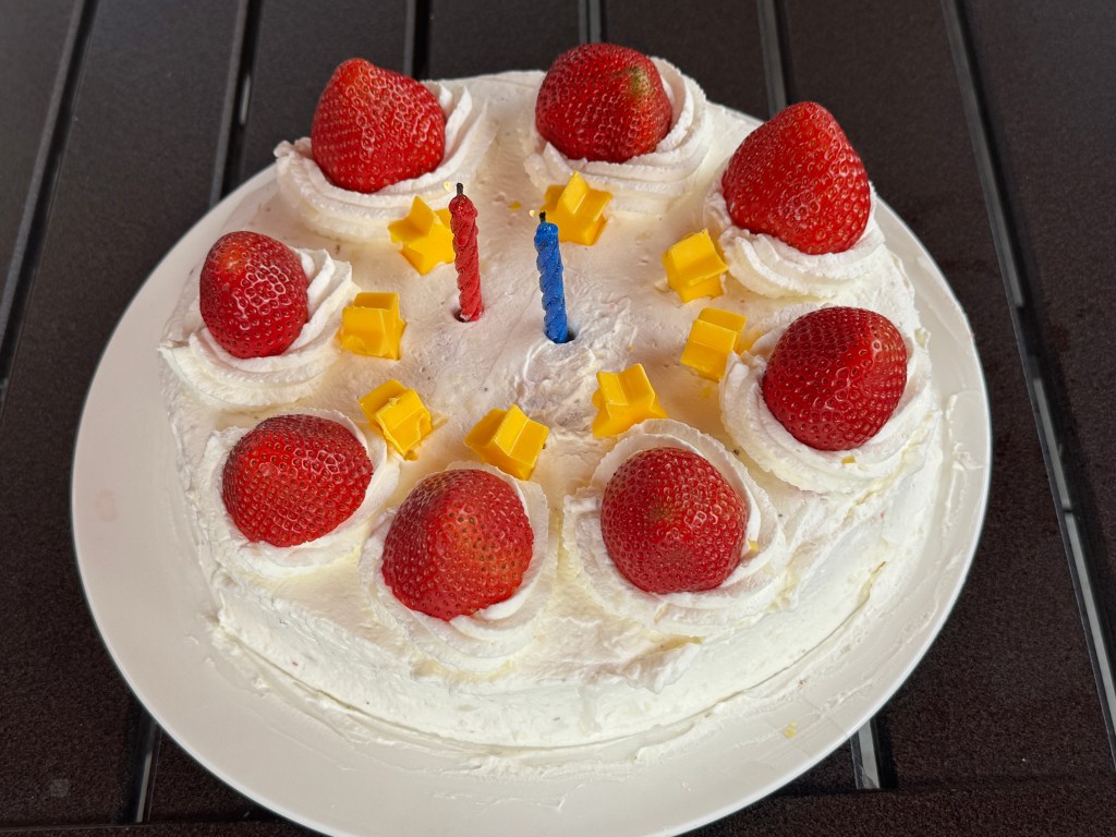Fraisier « Kirby » -- un gâteau rond et blanc, le glaçage en chantilly, avec une fraises sens dessus-dessous posées en haut, avec des étoiles jaunes. Il y a deux bougies au centre, une rouge et une bleue