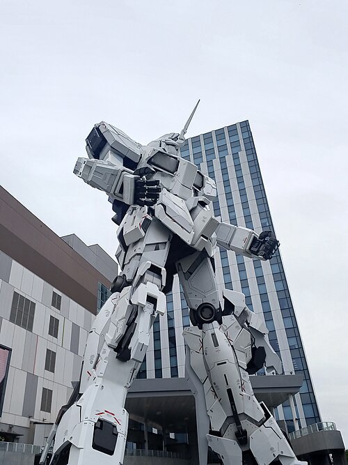 Photo d'aspect portrait qui montre un Gundam dans sa forme de robot plutôt qu'avion, à côté d'un gratte-ciel.