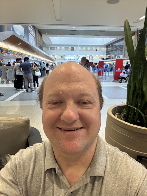 Moi dans un polo gris, avec la grande salle du terminal derrière moi. Il y a une plante vérité à ma gauche, et je ne regarde pas directement dans la photo car je me concentrais sur les contrôles pour la selfie. 