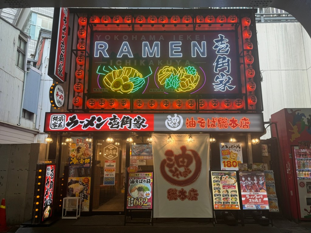 Il y a un panneau géant, presque la taille d'un étage avec le mot "RAMEN" en anglais, ainsi que les kanjis. Les lumières autour du panneau sont rouges. Il y a des bols de ramen en néons verts et marrons. Le premier étage, c'est le resto lui-même -- les prix sont bas (5-10 €), mais ça n'a pas l'air d'un endroit accueillant aux touristes.