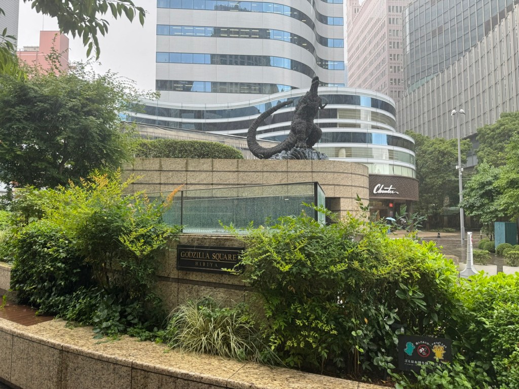 C'est un place recouverte de plantes vertes avec un panneau qui dit "Godzilla Square". La statue de Godzilla est en haut d'un bâtiment en pierre marron, et derrière ça, il y a un gratte-ciel largement en verre.