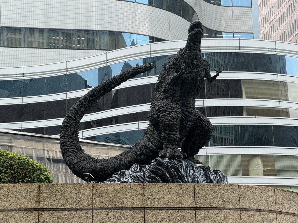 Vue de gauche de Godzilla. La statue est en métal noir, et il a une grosse queue. Il semble de faire trembler la terre -- le piédestal ressemble à une explosion.
