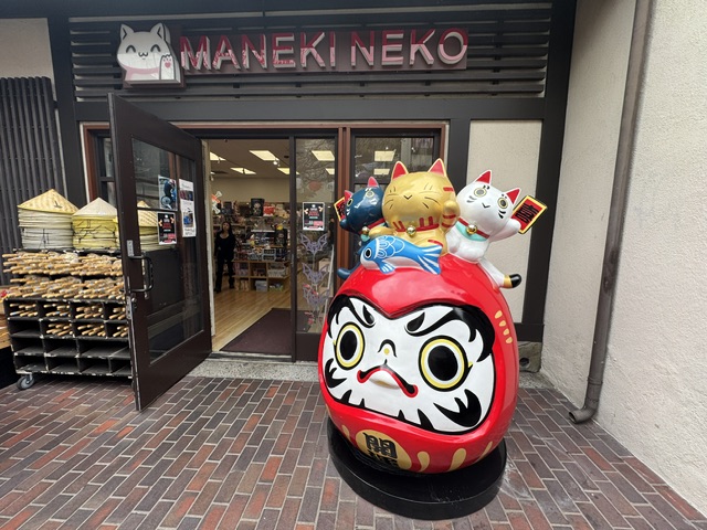 Entrée du magasin Maneki Neko. Devant la porte, il y a une statue rouge d'un chat, surmontée de deux petits « chats chanceux », doré et blanc