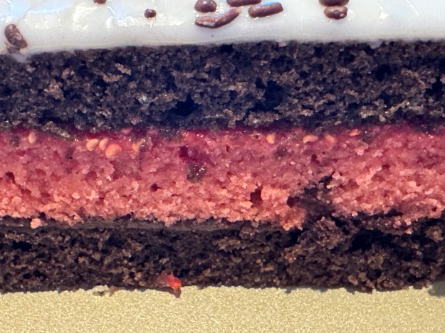 Vue en gros-plan du côté du gâteau. De bas en haut: couche de gâteau au chocolat, couche de ganache au chocolat, couche de gâteau à la framboise, couche de confiture de framboise, une autre couche de gâteau au chocolat, fondant blanc avec des vermicelles au chocolat.
