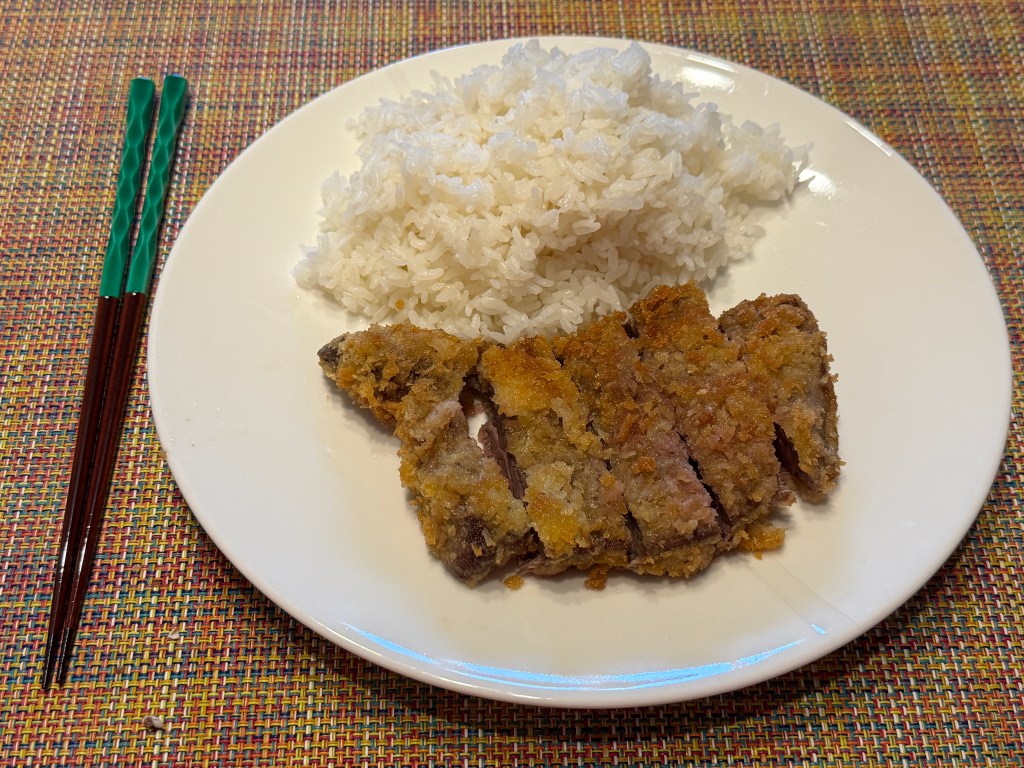 Steak pané et tranché vers le fond de l'assiette. En haut, il y a un tas de riz blanc. À gauche, il y a deux baguettes (l'ustensile, pas de pain), un bois bien marron avec des parties vertes en céramique vers le haut. Je les ai achetées au Japon.