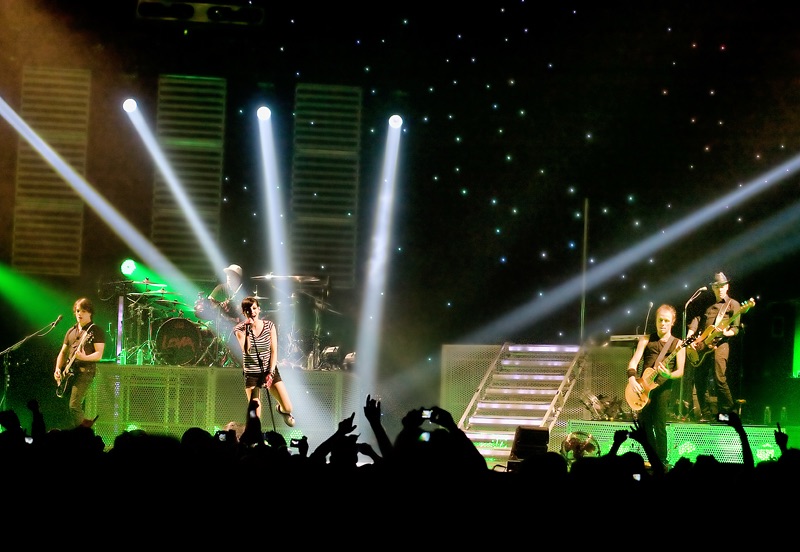 Le groupe Superbus sur scène. Le plateau est illuminé en vert, et il y a 5 projecteurs qui pointent tous à la chanteuse.