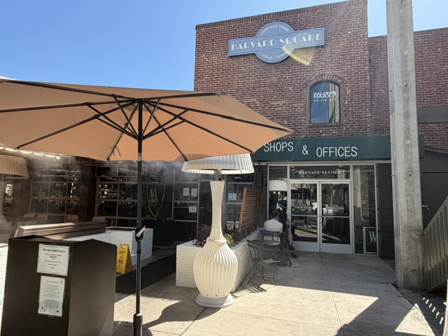 À gauche, il y a un parasol de table géant au-dessus d'une ardoise et un bureau de réception. Il y a un bâtiment de deux étages en brique, avec le nom "Harvard Square" visible sur un panneau noir. L'entrée du resto n'est pas bien visible.