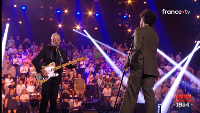 Michael Jones à gauche avec une guitare ; M. Gregorio à droite, tient un micro dans les mains