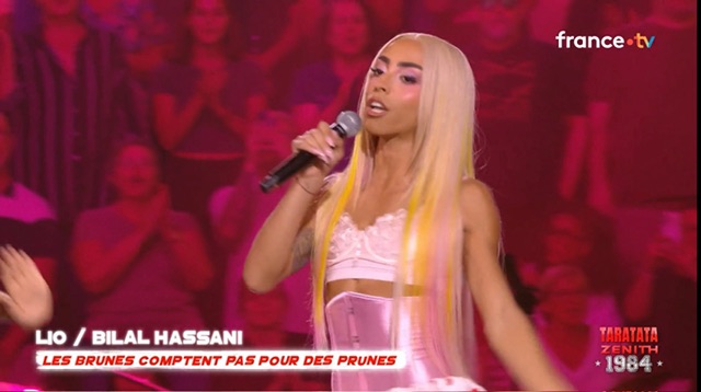M. Bilal Hassani avec une robe rose et des cheveux longs teints blonds pour ressembler à une femme. C'est du drag.