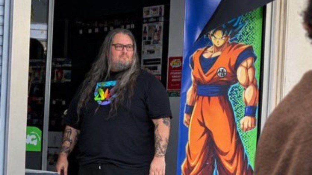 Pierre, un homme barbu avec des cheveux longs et en surpoids, reste debout dans l'entrée de son magasin, à côté de la porte, où un dessin de Goku s'affiche.